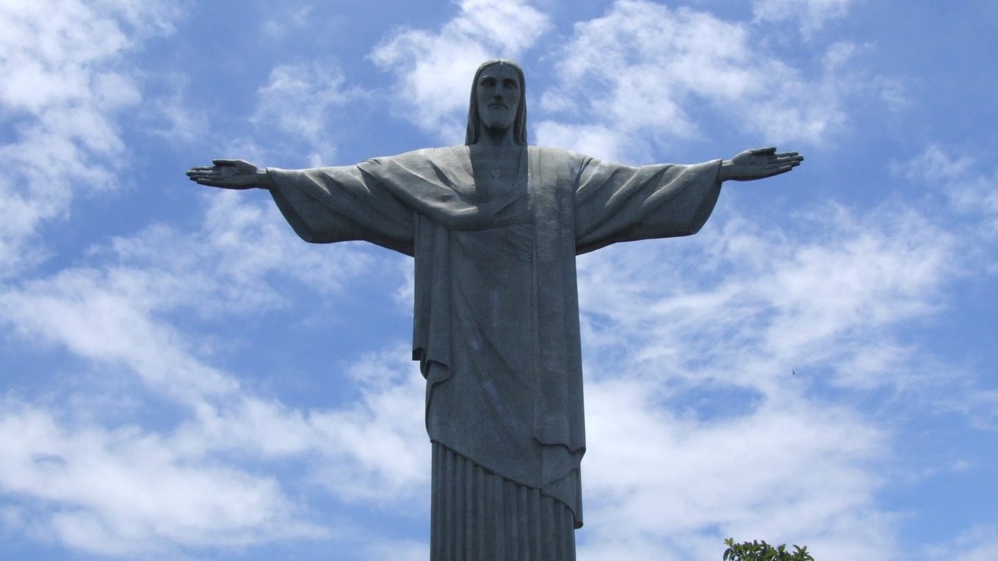 The Christ the Redeemer statue standing atop Corcovado mountain with its arms outstretched against a partly cloudy sky, overlooking the forested mountainside below.