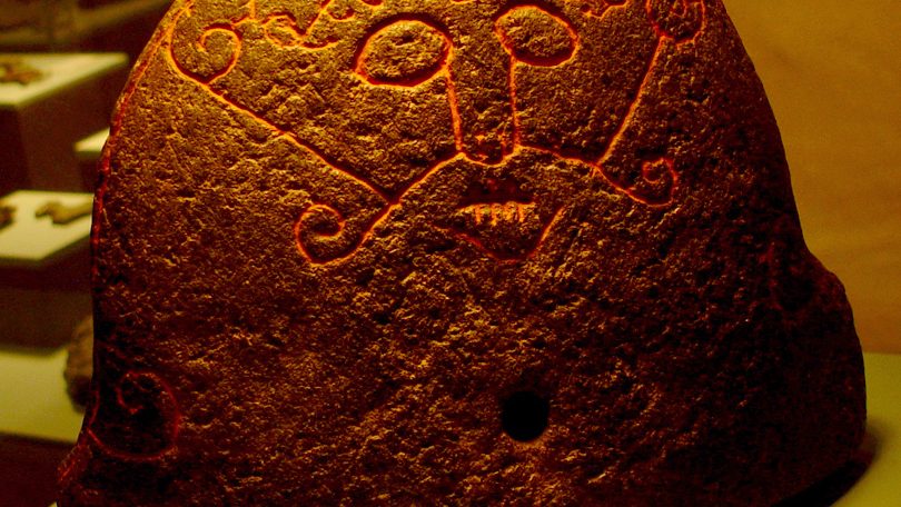 The Snaptun Stone, a curved hearth stone carved with a bearded face whose lips are stitched shut, in a museum case.